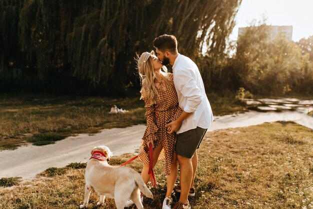 男と女は柳の背景にキスします。ロマンチックなカップルは彼らの最愛の犬と一緒に朝の散歩をしています。