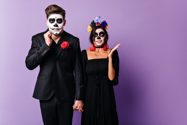 Guy and girl with painted faces are embarrassed, posing in purple studio. Man and woman hold hands.