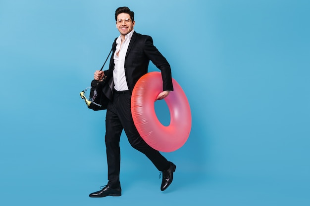 Free photo guy in glasses holds laptop bag. man in suit wants to go on vacation and poses with diving mask and pink inflatable circle.