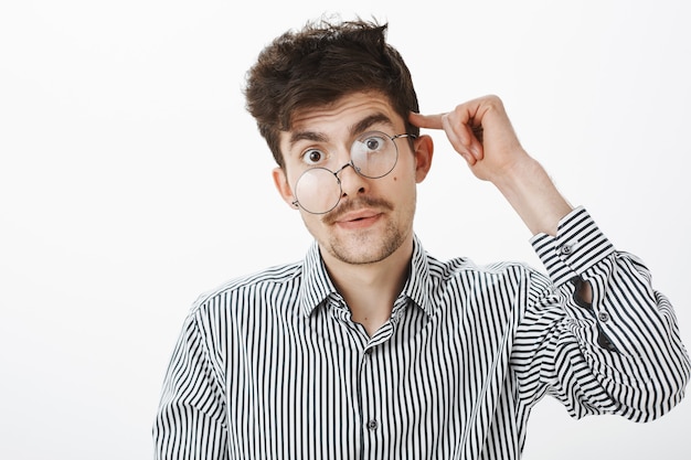 Guy goes crazy after night at work. Portrait of tired and stressed messy male model with beard and moustache, rolling index finger on temple, being confused and fed up, standing over gray wall