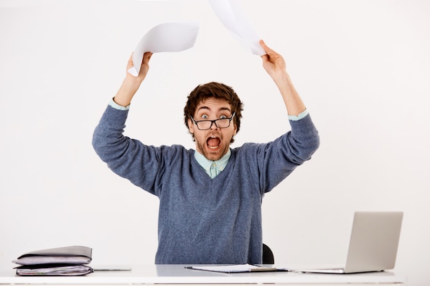 Guy going crazy with deadlines at work, tossing documents at reports, staring distressed and nervous, sit desk with laptop