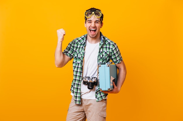 Guy in green shirt and beige shorts emotionally rejoices and clenches his fist. Man with diving mask, retro camera and suitcase laughs on orange space.