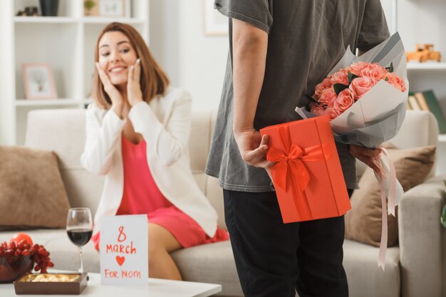 Guy holding present with bouquet smiling woman putting hands on cheeks sitting on sofa on happy women's day in living room