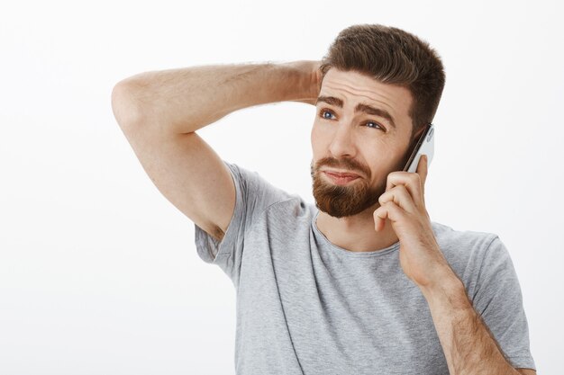 Guy intense, awkward trying say no during phone call. Unsure hesitating handsome boyfriend with beard and sick eyebrows scratching back of head gazing up holding cellphone near ear deciding how answer