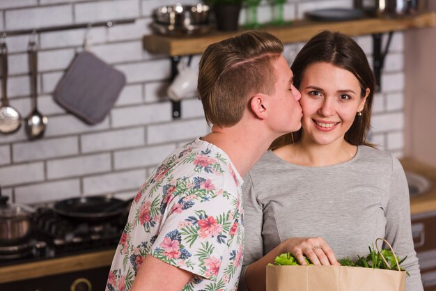 Guy kissing smiling girlfriend in kitchen