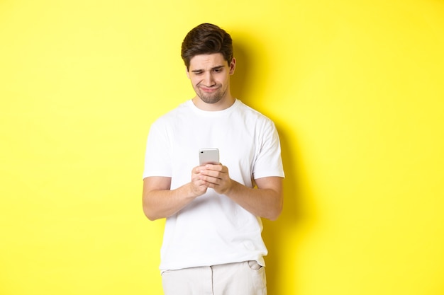 Guy looking displeased at smartphone screen, reading strange message on phone, standing in white t