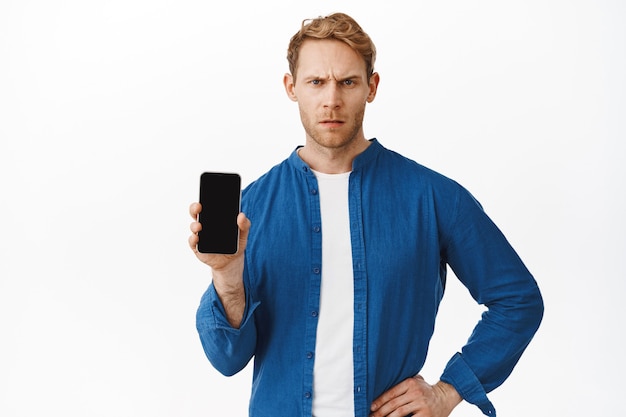 Guy looking frustrated while demonstrate something on mobile phone display, complaining, white wall