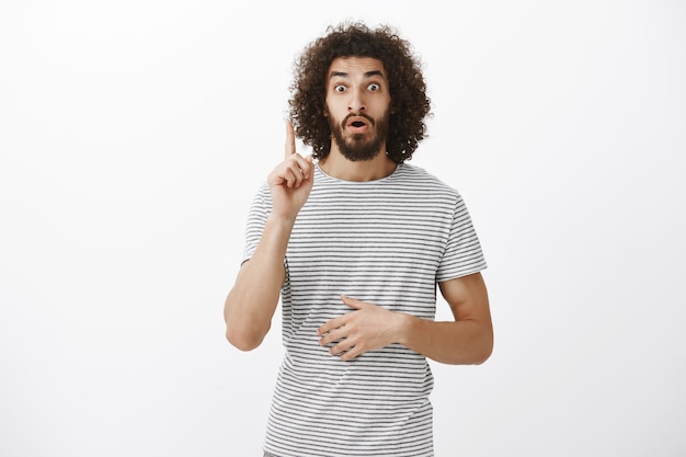 Guy made up great idea how to solve problem. Portrait of relieved surprised eastern guy with curly hair and beard raising index finger in eureka gesture