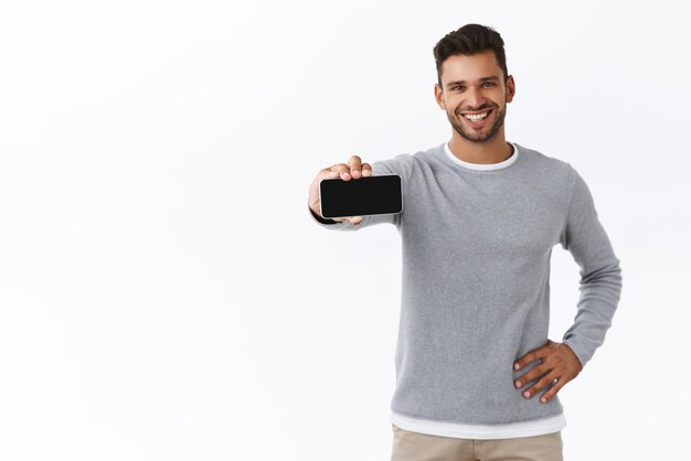 Guy offer download good app recommend gadget application Goodlooking sporty male in grey sweater pull smartphone at camera to show mobile screen horizontally smiling proud and pleased