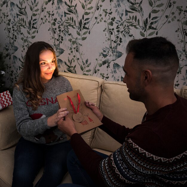 Guy presenting gift packet to cheerful lady on settee