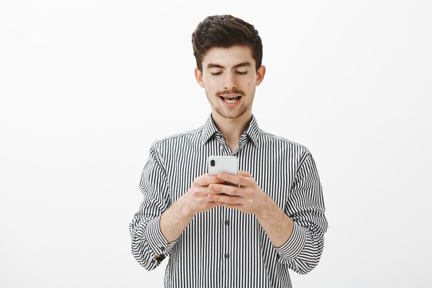Guy reading out loud message he received on phone. Portrait of carefree handsome young man with beard and moustache, typing in smartphone, looking at screen focused and interested over gray wall