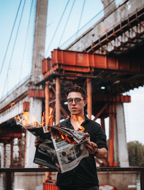 Guy reads a burning newspaper in front of a bridge
