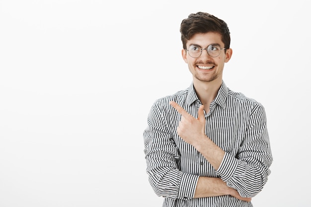 Guy recalled funny moment. Portrait of interested good-looking european male freelancer in glasses, pointing at upper left corner and smiling broadly, thinking about joke, standing over gray wall