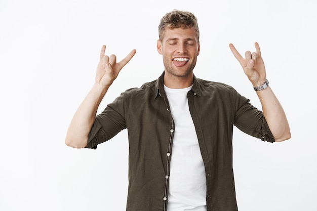 Guy rocking, feeling cool and awesome showing rock-n-roll gesture close eyes and stick tongue excited and carefree feeling happy lifting up mood from awesome vibes after concert over gray wall