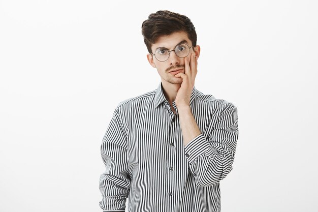 Guy shocked how stupid his coworker is. Portrait of stunned caucasian male model with moustache, touching cheek and staring with clueless expression, being shocked with dumb person