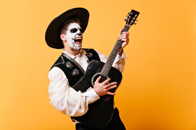 감정적 인 노래를 부르는 남자는 기타를 연주합니다. 솜브레로에 그려진 얼굴을 가진 남자의 초상화,