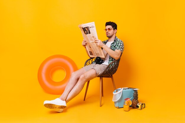 Guy sits and reads newspaper on space of suitcase, pineapple and rubber ring.