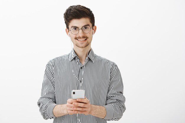 Guy taking notes while listening audiobook. Portrait of friendly attractive caucasian man with moustache and beard, wearing earphones, enjoying watching great video in smartphone, smiling