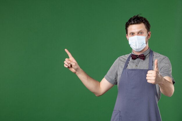 guy waiter in uniform with medical mask and pointing up on the right side making ok gesture on green background