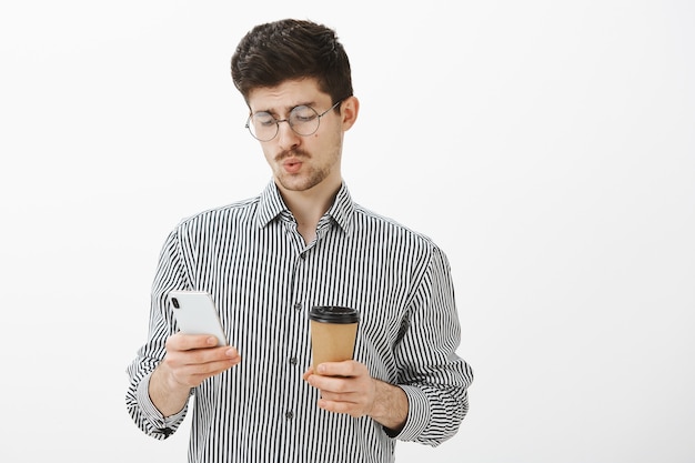 ボスからの厄介なメッセージを見て、口笛を吹く男。ひげと口ひげのアイウェア、コーヒーを飲み、スマートフォンでスクロールフィードと無関心なハンサムなヨーロッパの男性モデルの肖像画