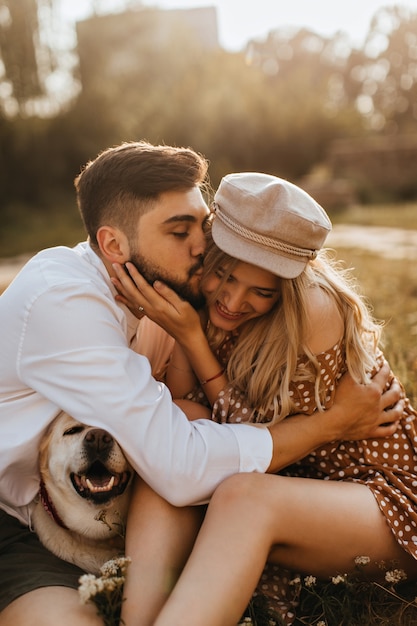 Guy in white shirt hugs and kisses his beloved girlfriend on cheek. Woman in polka-dot dress and her husband fooling around with their dog in park.