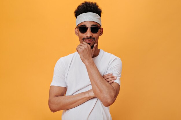 Guy in white T-shirt and headband looks at camera on orange wall