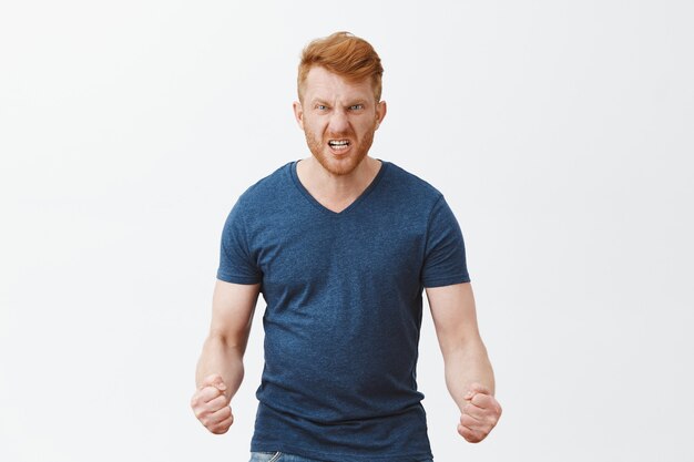 Guy will show how anger looks. Portrait of pissed and angry dangerous redhead guy with outraged expression, wrinkling nose, frowning and clenching fists dangerously, feeling hate over gray wall