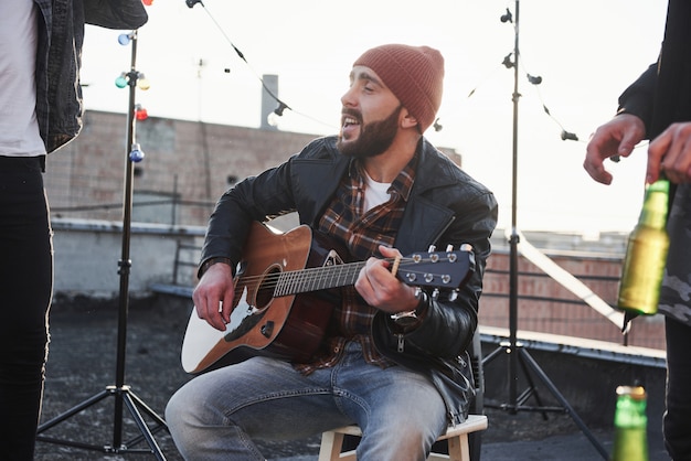 Ragazzo con chitarra acustica canta. gli amici si divertono alla festa sul tetto con lampadine colorate decorative