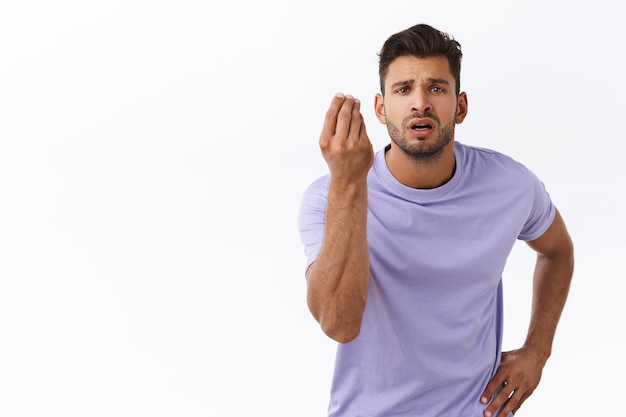 Foto gratuita ragazzo con la barba in maglietta viola che agita le dita in un gesto italiano, dove sono i miei soldi o cosa vuoi, accigliato infastidito, in piedi interrogato e deluso