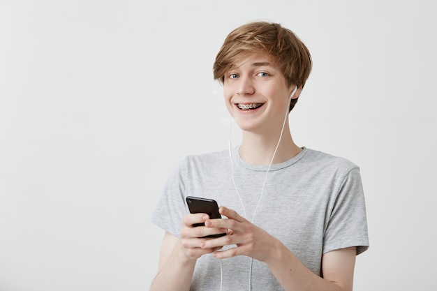 Guy with fair hair, wearing gray t-shirt holding modern smart phone using high-speed Internet connection, texting messages to his friends, smiling broadly. Modern technology and communication.