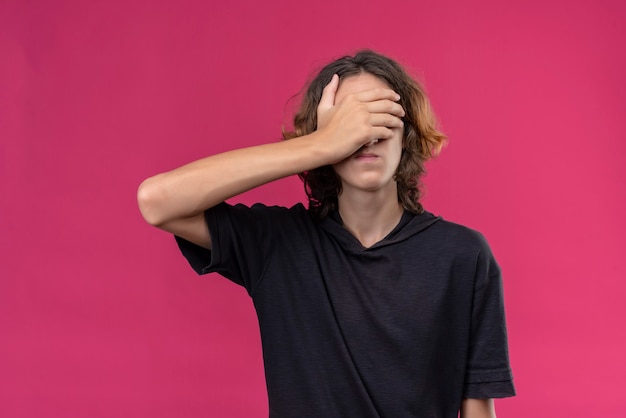 Парень с длинными волосами в черной футболке закрыл лицо рукой на розовой стене