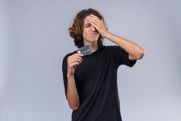 Guy with long hair in black t-shirt holding a bank card and grabbed his head on white wall