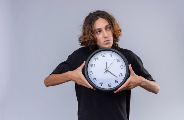 Парень с длинными волосами в черной футболке держит настенные часы на белой стене