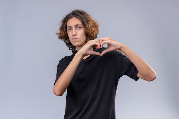 Ragazzo con i capelli lunghi in maglietta nera mostra il cuore con le mani sul muro bianco