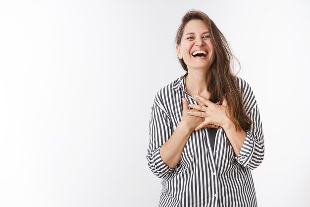 Ha-ha so amusing. Portrait of charming carefree middle-aged woman in striped blouse holding hands on chest touched and pleased laughing out loud amazed and entertained standing over white wall