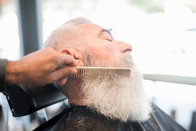 Hairdresser combing beard of aged client in barbershop