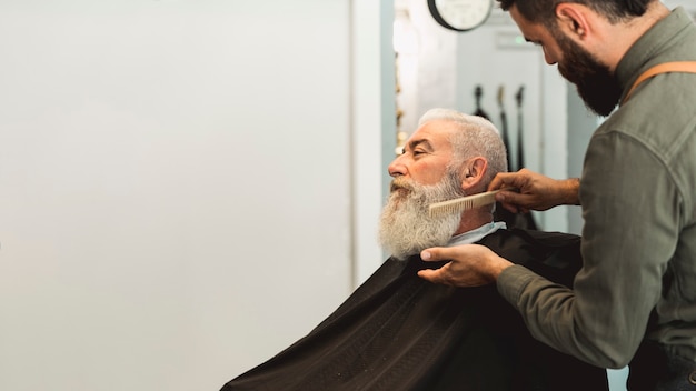 Foto gratuita parrucchiere che pettina barba al cliente senior in salone