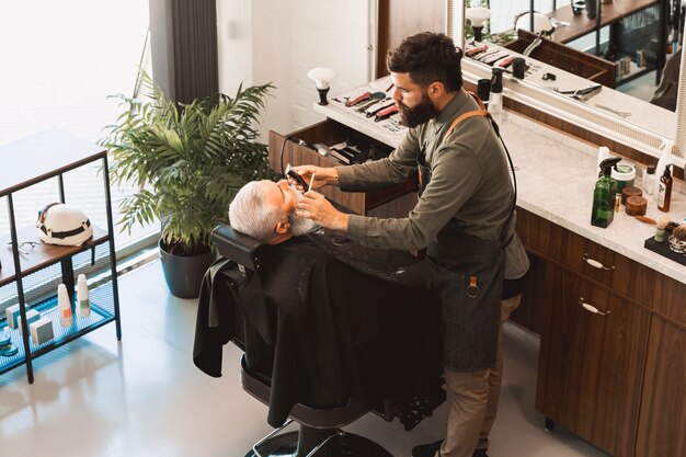 Парикмахер выпрямляет бороду бритвой и расческой старшего клиента