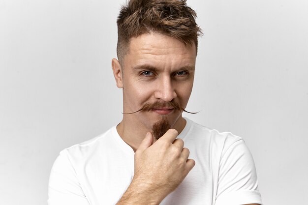 Hairdressing, fashion and style concept. Picture of trendy looking man with stylish haircut, mustache and goatee beard staring at camera with mysterious smile, looking from under his thick eyebrows