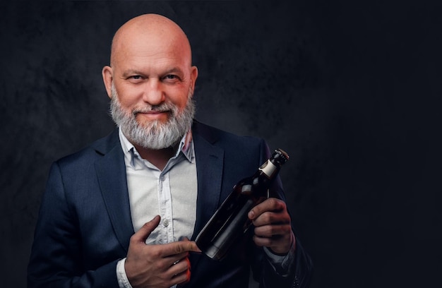 Hairless elderly man dressed in stylish suit holding bottle against dark background.