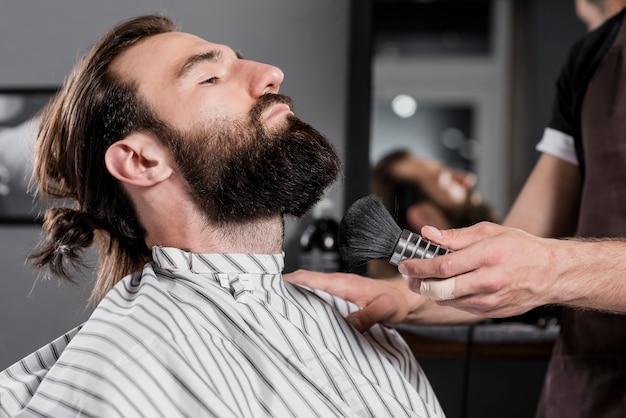 Hairstylist holding shaving brush near man's bearded face