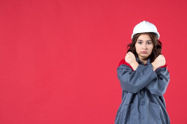 Half body shot of angry female builder in uniform with hard hat and making stop gesture on isolated red background