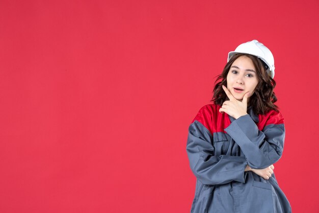 Half body shot of confident female builder in uniform with hard hat and concentrated on something on isolated red background