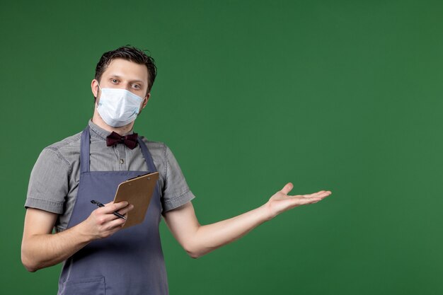 Half body shot of confident guy server in uniform with medical mask and holding check book pen pointing something on the left side on green background