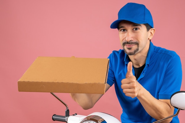 Half body shot of confident male delivery person wearing hat sitting on scooter holding order making ok gesture