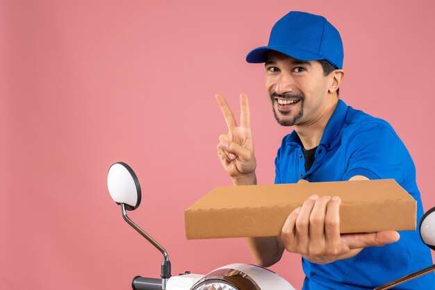 Half body shot of confident male delivery person wearing hat sitting on scooter holding order making victory gesture