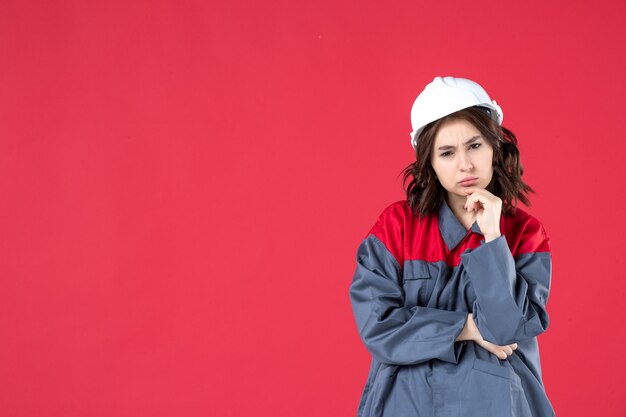 Half body shot of questioning female builder in uniform with hard hat and concentrated on something on isolated red background