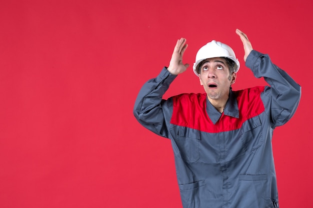 Half body shot of scared young architect in uniform with hard hat and looking up on isolated red wall