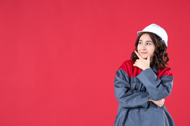 Снимок половины тела улыбающейся женщины-строителя в униформе с каской, сосредоточенной на чем-то на изолированном красном фоне