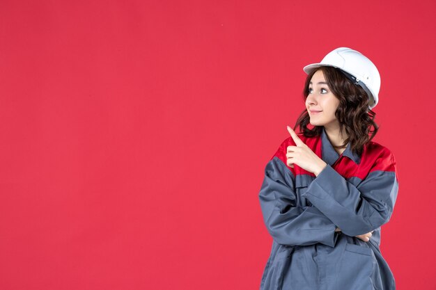 Half body shot of smiling focused female builder in uniform with hard hat and pointing up on the right side on isolated red background